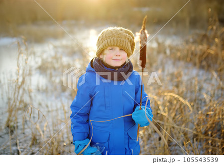 Cute preschool boy is playing on the ice of a frozen lake or river on a cold sunny winter sunset. Child having fun with icicle and dry reed plant during family hiking. Kids outdoor games Cute preschool boy is playing on the ice of a frozen lake or river on a cold sunny winter sunset. Child having fun with icicle and dry reed plant during family hiking. Kids outdoor games 105543539
