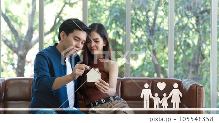 Young asian couple sits on a cozy couch, putting silver coin into a wooden house-shaped storing coin box , hinting at dreams of an idyllic future. Young asian couple sits on a cozy couch, putting silver coin into a wooden house-shaped storing coin box , hinting at dreams of an idyllic future. 105548358
