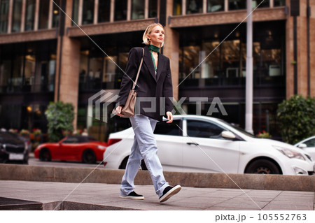 Beautiful young stylish woman walking down city street against backdrop of building, wearing fashionable clothes jeans, black blazer, handbag and sneakers. Fashion model serious and looks away 105552763