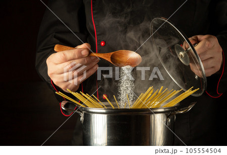 The cook adds a spoonful of salt to a boiling pot of spaghetti. Free space for recipe or menu on dark background. The cook adds a spoonful of salt to a boiling pot of spaghetti. Free space for recipe or menu on dark background. 105554504