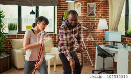 Diverse life partners dancing and sweeping dust off floors, using vacuum cleaner and washing solution. Cheerful couple laughing and enjoying spring cleaning in apartment, household chores. 105556310
