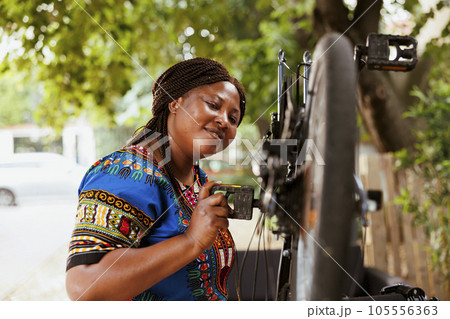 Active sporty african american woman maintains and repairs bicycle parts outside using specialized tools. Female cyclist with enthusiasm meticulously inspecting and repairing damaged bicycle. 105556363
