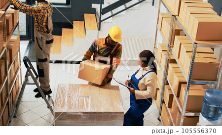 Group of people working on stock logistics in depot, organizing pallets of boxes on racks and shelves. Team of employees moving cardboard boxes in storage room. Handheld shot. Group of people working on stock logistics in depot, organizing pallets of boxes on racks and shelves. Team of employees moving cardboard boxes in storage room. Handheld shot. 105556370