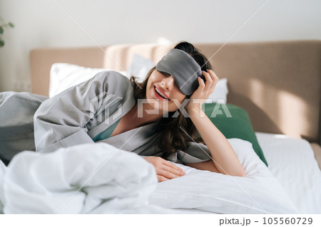 Portrait of cheerful early bird female wearing sleeping mask lying in comfy bed in luxury hotel bedroom looking at camera. Charming brunette young woman in pajama waking up enjoy good morning at home. 105560729