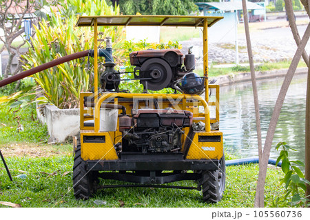 Sprinkler truck small old yellow water pump is watering trees in park. with old engine that works well. Parked on green grass by pond. By connecting pipe to pump water help water plants in dry season. 105560736