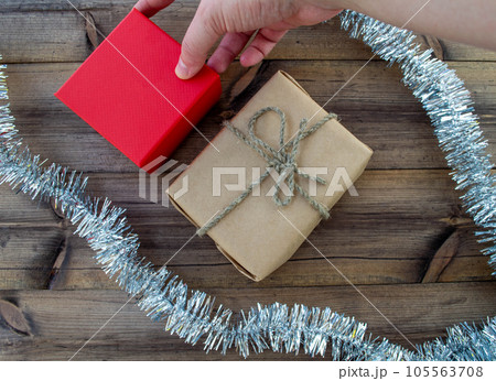 New Year gifts. A gift in craft paper, a hand holds a gift in a red box, tinsel on a wooden background. 105563708
