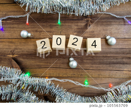 New Year's card 2024. Wooden cubes with numbers 2024, tinsel, garland and New Year's balls on a wooden background. New Year's card 2024. Wooden cubes with numbers 2024, tinsel, garland and New Year's balls on a wooden background. 105563709
