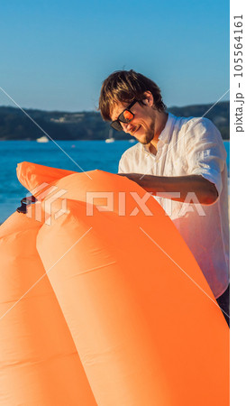 Summer lifestyle portrait of man inflates an inflatable orange sofa on the beach of tropical island. Relaxing and enjoying life on air bed VERTICAL FORMAT for Instagram mobile story or stories size 105564161