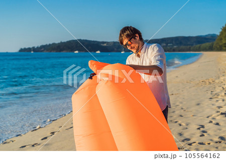 Summer lifestyle portrait of man inflates an inflatable orange sofa on the beach of tropical island. Relaxing and enjoying life on air bed 105564162