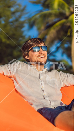 Summer lifestyle portrait of man sitting on the orange inflatable sofa on the beach of tropical island. Relaxing and enjoying life on air bed VERTICAL FORMAT for Instagram mobile story or stories size 105564238