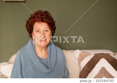 Portrait of an elderly woman with blue eyes and red hair looks at the camera in a room sitting on a sofa 105564785