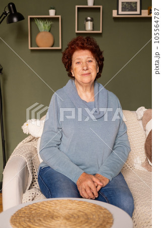Portrait of an elderly woman with blue eyes and red hair looks at the camera in a room sitting on a sofa 105564787