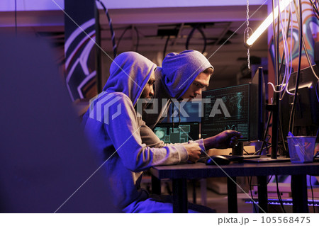 Hacker demonstrating malicious software on computer to illegal malware developer. Diverse criminals in hoods cracking strong password and using ransomware in abandoned warehouse Hacker demonstrating malicious software on computer to illegal malware developer. Diverse criminals in hoods cracking strong password and using ransomware in abandoned warehouse 105568475