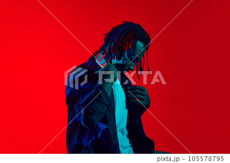 Musician. Handsome, stylish young guy with dreads posing against red background in neon light 105578795