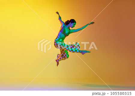 Young woman, professional contemporary dancer in motion, in stylish clothes against gradient yellow orange background in neon light 105580315