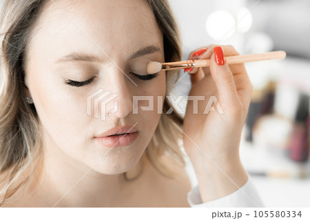 A young woman closes her eyes when makeup artist neatly applies different shades shadow to her eyelids, creating magical effect depth and accentuating her look. Makeup in beauty salon on wedding day A young woman closes her eyes when makeup artist neatly applies different shades shadow to her eyelids, creating magical effect depth and accentuating her look. Makeup in beauty salon on wedding day 105580334
