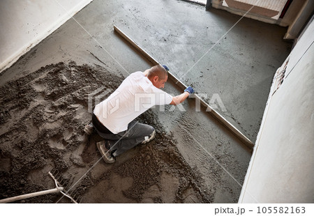 Man smoothing and leveling surface with straight edge while screeding floor in apartment. Top view of male construction worker placing screed rail on the floor covered with sand-cement mix. Man smoothing and leveling surface with straight edge while screeding floor in apartment. Top view of male construction worker placing screed rail on the floor covered with sand-cement mix. 105582163