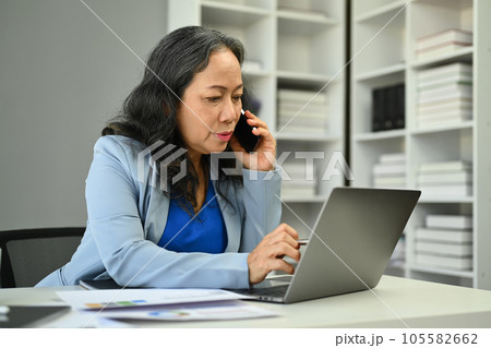 Professional 50s businesswoman having phone conversation and using laptop at desk. 105582662
