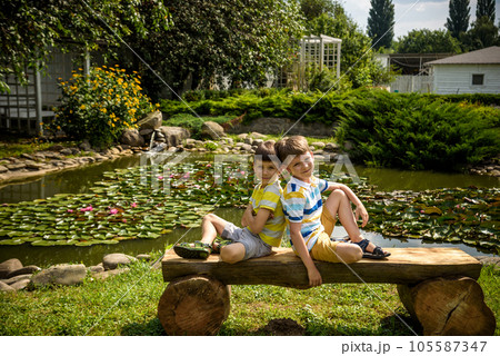 Two brother boys sitting on a bench in a park near peaceful lake with water lily Nymphaeum. Kids relaxing on nature on hot summer day Two brother boys sitting on a bench in a park near peaceful lake with water lily Nymphaeum. Kids relaxing on nature on hot summer day 105587347
