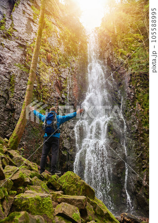 Tourist attraction of Germany - falls of Burgbach Waterfall near Schapbach, Black Forest, Baden-Wurttemberg, Germany. Man hiker in blue jacket standing on stone and looks at flow of falling water 105588995