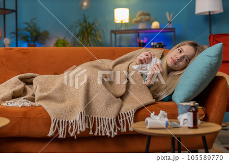 Sick ill young woman suffering from cold, allergy, lying on home sofa sneezes wipes snot into napkin Sick ill young woman suffering from cold, allergy, lying on home sofa sneezes wipes snot into napkin 105589770