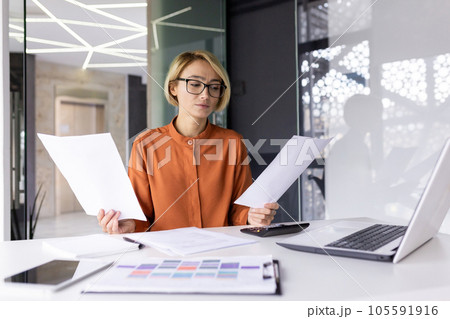 Serious and pensive business woman behind paper work inside office, female financier worker thinks about contracts and reports with charts and graphs, blonde successful woman uses laptop at work. Serious and pensive business woman behind paper work inside office, female financier worker thinks about contracts and reports with charts and graphs, blonde successful woman uses laptop at work. 105591916