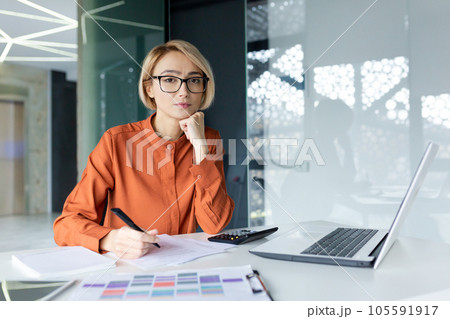 Portrait of young thinking business woman, blonde at workplace inside office concentrating and looking seriously at camera, female boss using laptop behind paper work with documents and reports. Portrait of young thinking business woman, blonde at workplace inside office concentrating and looking seriously at camera, female boss using laptop behind paper work with documents and reports. 105591917