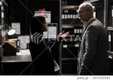 Accountancy asian assistant reporting budget plan optimizations to elderly businessman executive manager. Dark file cabinet office room surrounded by document folders on cabinet shelves Accountancy asian assistant reporting budget plan optimizations to elderly businessman executive manager. Dark file cabinet office room surrounded by document folders on cabinet shelves 105592892