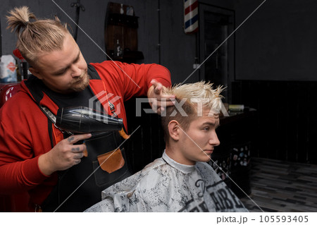 Stylish professional European man barber blow-drying hair for a client guy in a haircut in a barbershop 105593405