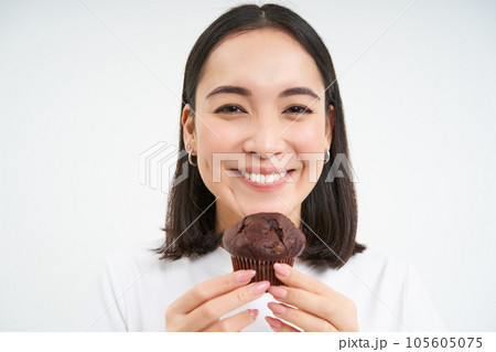 Pastry, sweets and bakery. Close up of smiling asian woman, biting cupcake with joy, standing isolated on white background 105605075