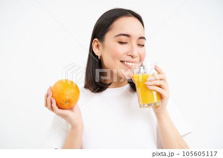 Close up portrait of asian woman, drinking glass of orange juice with delight and happy smile, white studio background 105605117