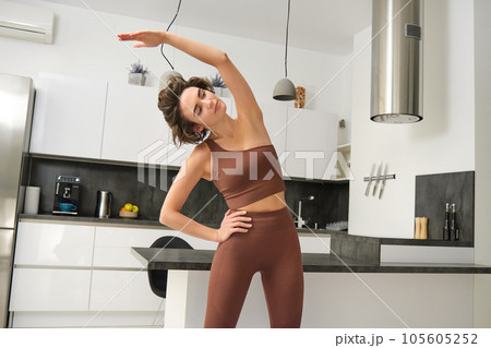 Home workout concept. Young woman in fitness clothes, doing stretching exercises, training at home behind kitchen, pulling arm to one side, focusing on body 105605252