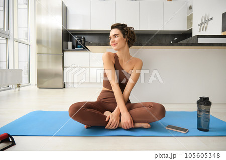 Portrait of fitness girl doing yoga on rubber mat at home, workout indoors in kitchen wearing sportswear, practice minfulness, sitting in lotus pose with water bottle next to her 105605348