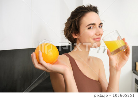 Image of sportswoman, fitness girl holding glass of juice and an orange, smiling, drinking vitamin beverage after workout, standing in her kitchen at home. Healthy lifestyle and sport concept Image of sportswoman, fitness girl holding glass of juice and an orange, smiling, drinking vitamin beverage after workout, standing in her kitchen at home. Healthy lifestyle and sport concept 105605551