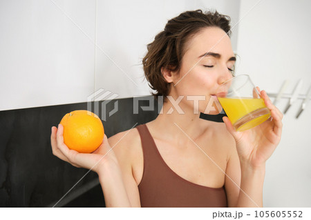 Healthy lifestyle and sport. Beautiful smiling woman, drinking fresh orange juice and holding fruit in her hand, enjoying her vitamin drink after workout at home, standing in kitchen Healthy lifestyle and sport. Beautiful smiling woman, drinking fresh orange juice and holding fruit in her hand, enjoying her vitamin drink after workout at home, standing in kitchen 105605552