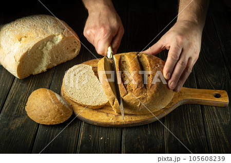 The cook cuts wheat bread with a knife on a cutting wooden board. Healthy food and traditional bakery concept on dark background The cook cuts wheat bread with a knife on a cutting wooden board. Healthy food and traditional bakery concept on dark background 105608239