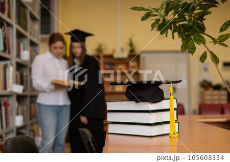 Close-up of a graduation cap on a stack of books in a library. teacher and young female graduate in the background. Close-up of a graduation cap on a stack of books in a library. teacher and young female graduate in the background. 105608334