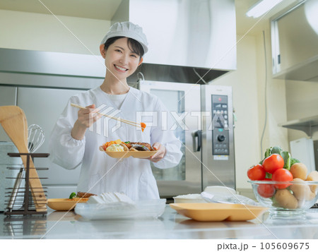 調理室でお弁当におかずを詰める調理服を着た若い女性 調理室でお弁当におかずを詰める調理服を着た若い女性 105609675