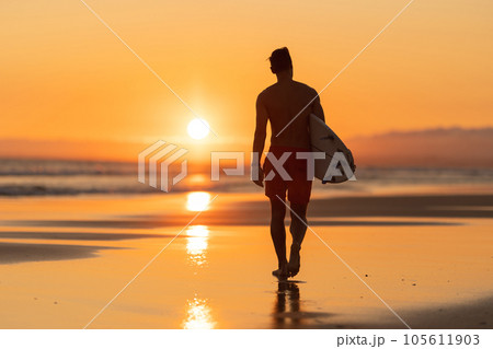 Black silhouette of an attractive man walking on the shore holding a surfboard at orange sunset Black silhouette of an attractive man walking on the shore holding a surfboard at orange sunset 105611903