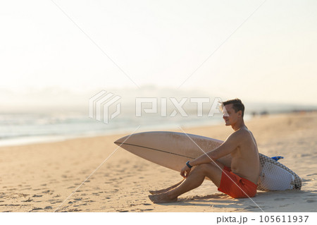 An attractive man with nice athletic body sitting on the shore holding a surfboard An attractive man with nice athletic body sitting on the shore holding a surfboard 105611937