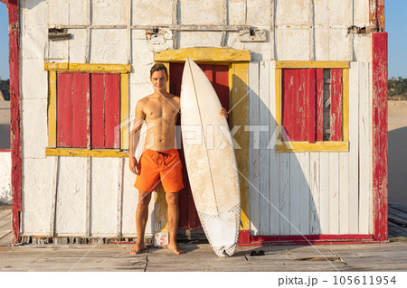 An attractive smiling man with nice athletic body standing at the old seaside house holding a surfboard 105611954