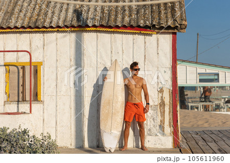 A man with nice athletic body wearing sunglasses standing at the shore house on the beach with his surfboard A man with nice athletic body wearing sunglasses standing at the shore house on the beach with his surfboard 105611960