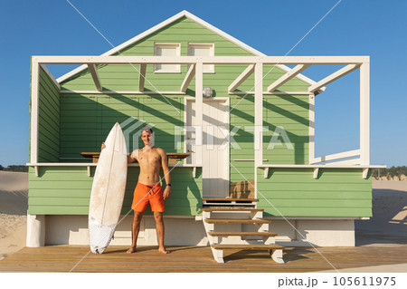 A smiling man surfer standing at a shore house on the beach holding his surfing board A smiling man surfer standing at a shore house on the beach holding his surfing board 105611975