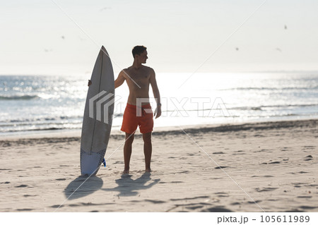 A shirtless man surfer standing on the beach holding a surfboard A shirtless man surfer standing on the beach holding a surfboard 105611989