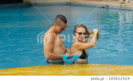 The loving couple hugs and kisses, drinking blue cocktail alcohol liquor in swimming pool at hotel outdoor. Portrait of caucasian man and woman. The loving couple hugs and kisses, drinking blue cocktail alcohol liquor in swimming pool at hotel outdoor. Portrait of caucasian man and woman. 105612424