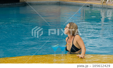 The wide middle shot pretty woman drinking blue cocktail alcohol liquor in swimming pool at hotel. Portrait of sexy girl in pool outdoor. The wide middle shot pretty woman drinking blue cocktail alcohol liquor in swimming pool at hotel. Portrait of sexy girl in pool outdoor. 105612429