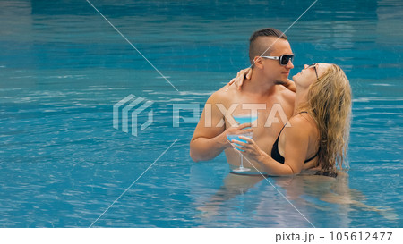 The loving couple hugs and kisses, drinking blue cocktail alcohol liquor in swimming pool at hotel outdoor. Portrait of caucasian man and woman. 105612477