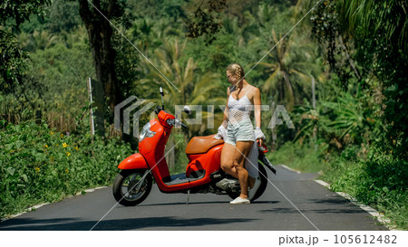 Woman on red scooter in white clothes drive on forest road trail. Dancing trip. One girl caucasian tourist in sunglasses dance, relax near motorbike. 105612482