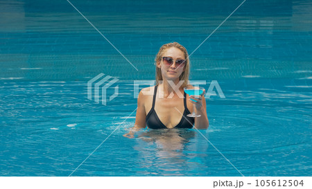 The wide middle shot pretty woman drinking blue cocktail alcohol liquor in swimming pool at hotel. Portrait of sexy girl in pool outdoor. The wide middle shot pretty woman drinking blue cocktail alcohol liquor in swimming pool at hotel. Portrait of sexy girl in pool outdoor. 105612504