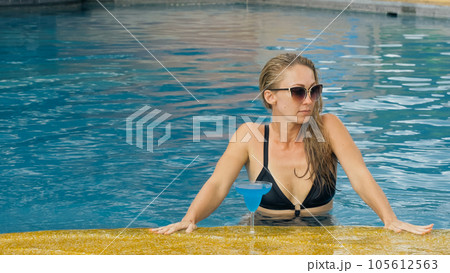 The wide middle shot pretty woman drinking blue cocktail alcohol liquor in swimming pool at hotel. Portrait of sexy girl in pool outdoor. The wide middle shot pretty woman drinking blue cocktail alcohol liquor in swimming pool at hotel. Portrait of sexy girl in pool outdoor. 105612563